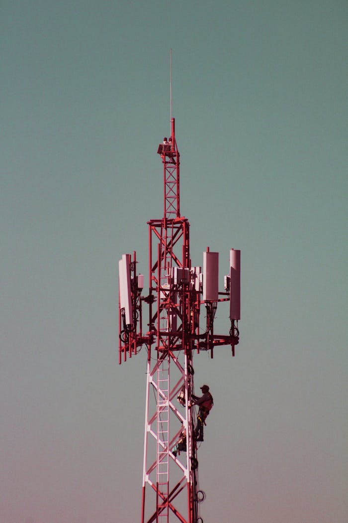 An adult male worker climbs a red communication tower against a clear sky, showcasing occupational safety and telecom infrastructure.